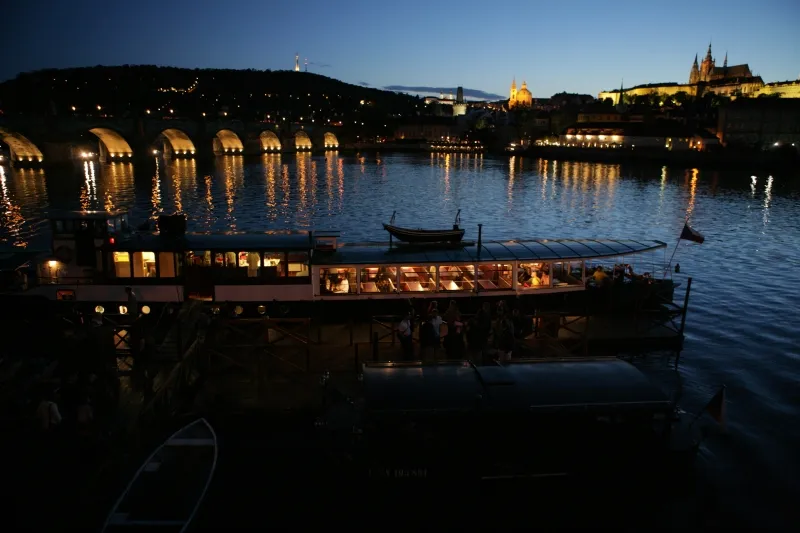 Karelsbrug bij schemering met verlichte bogen weerspiegeld in de rivier, een riviercruisboot op de voorgrond en de skyline van Praag op de achtergrond