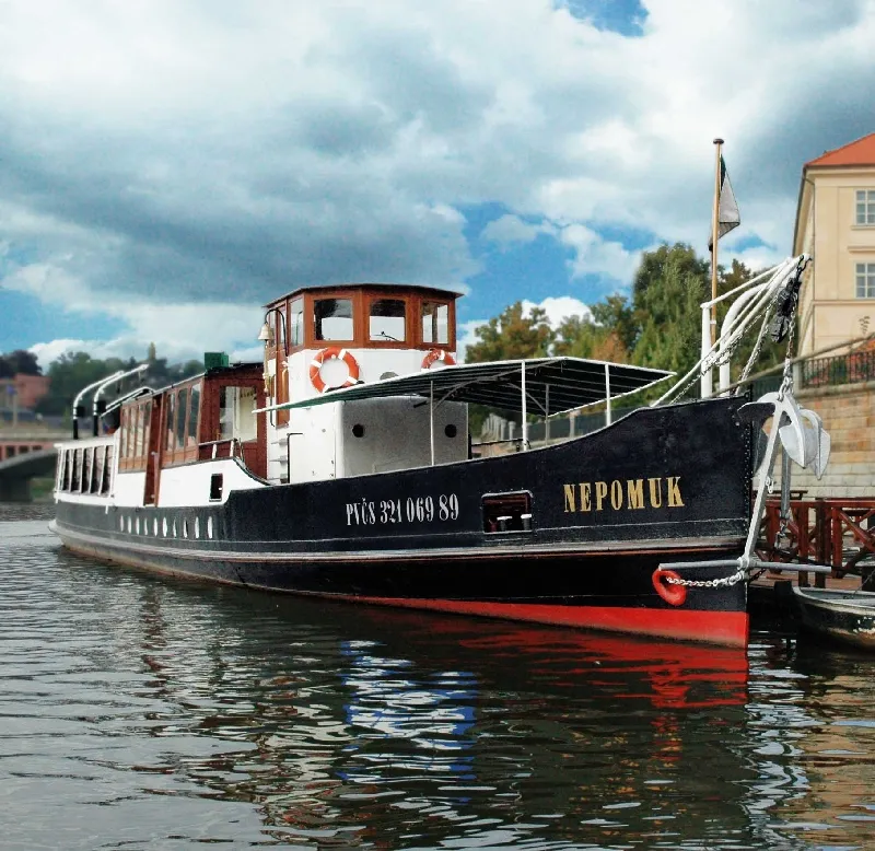 Een zwart-wit rivierboot genaamd NEPOMUK aangemeerd aan de Moldau met een brug en gebouwen op de achtergrond