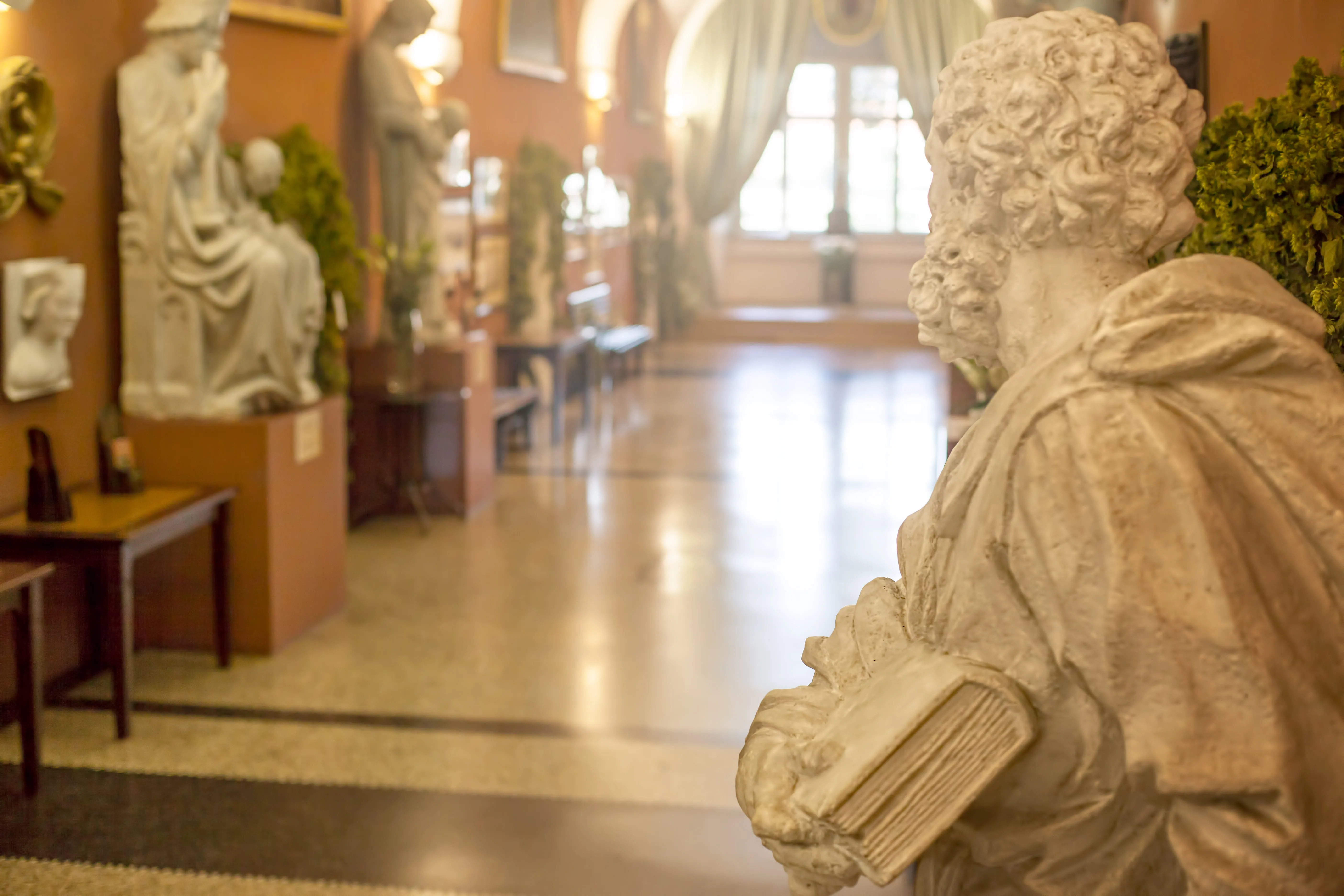 Een marmeren beeld van een geklede figuur met een boek op de voorgrond van een museumgalerij, met andere sculpturen en exposities op de achtergrond.