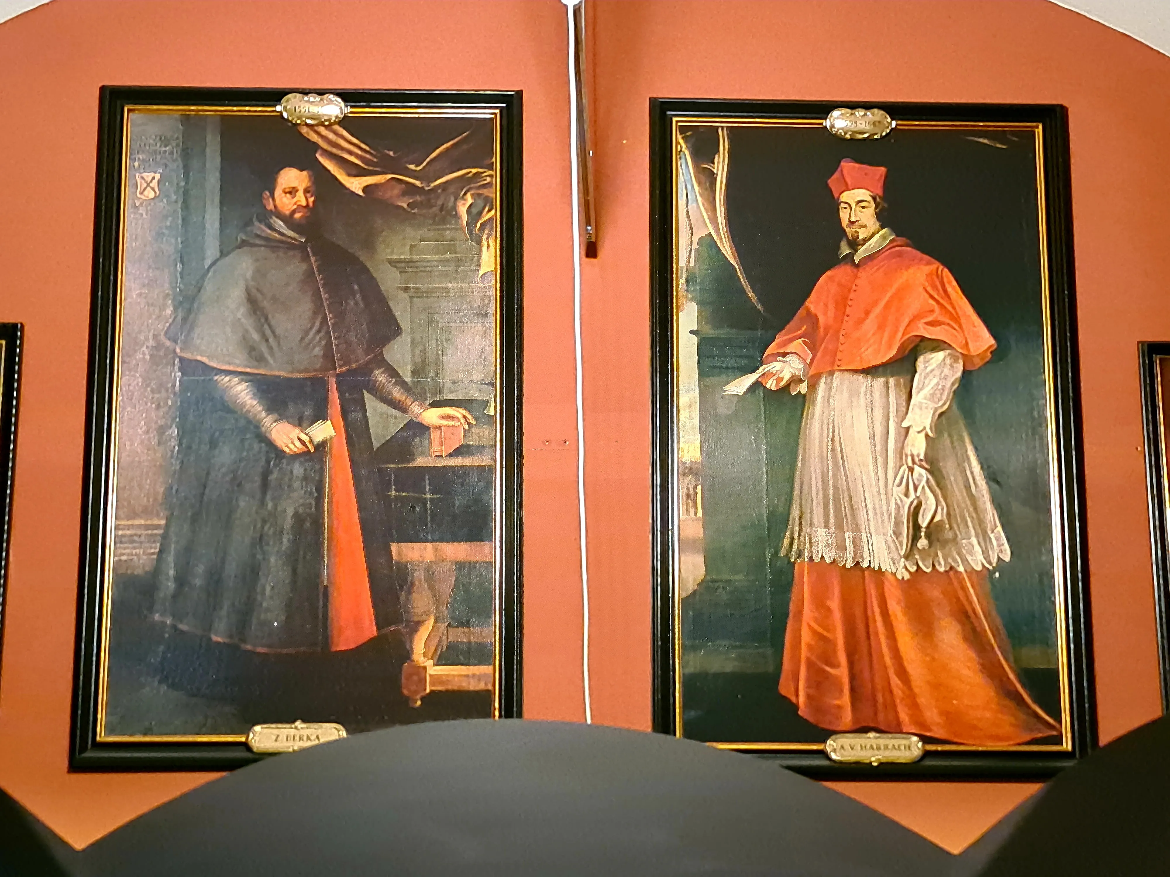 Twee ingelijste historische portretten aan een muur, waarvan één van een man in een donkere robe en de ander van een man in rood en wit religieus gewaad