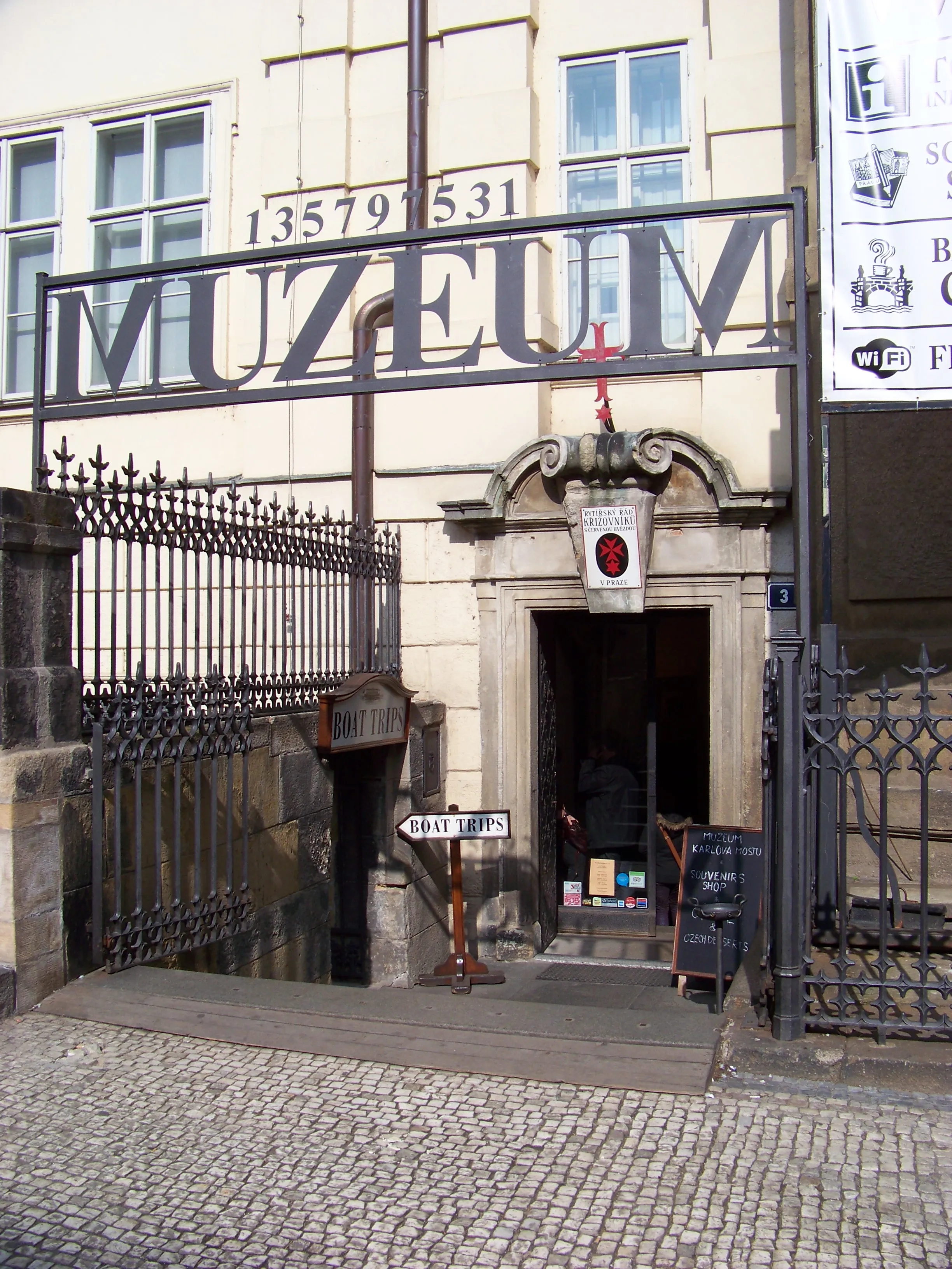 Ingang naar het Charles Bridge Museum met een groot 'MUZEUM'-bord boven de deur en een kasseienstraat ervoor