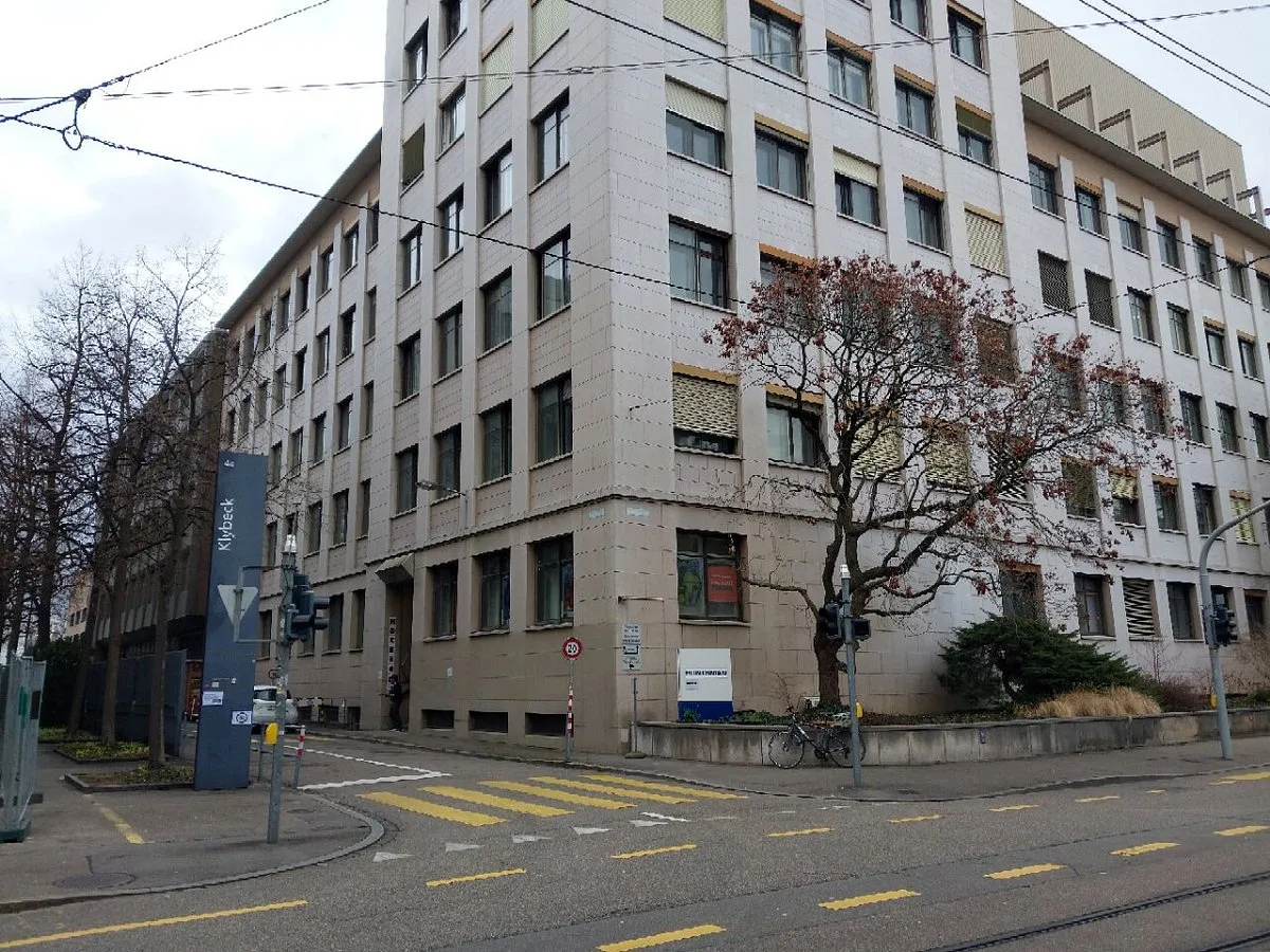 Vista de la esquina de un edificio beige de varias plantas con líneas de tranvía por encima y un letrero azul cerca de la entrada