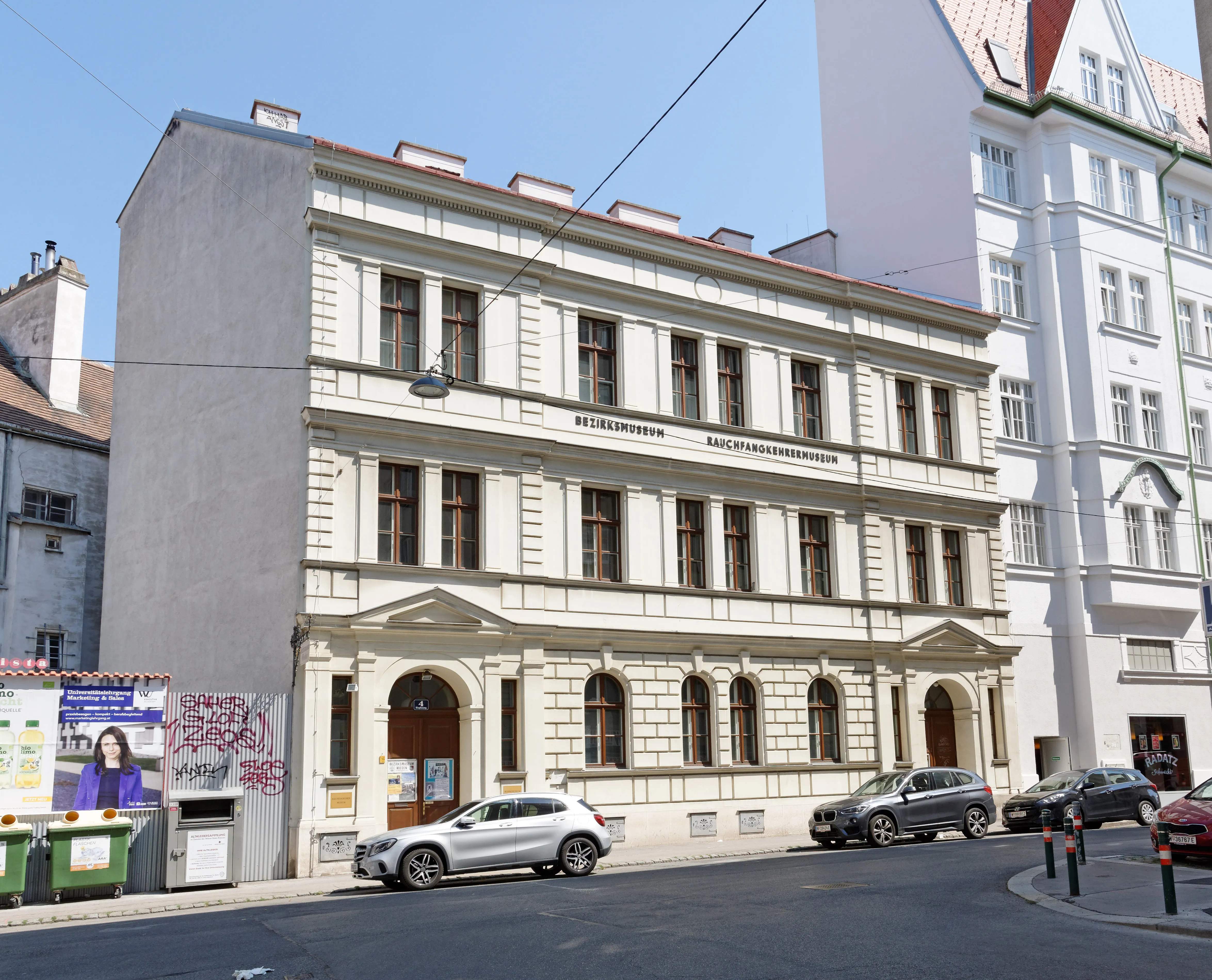 Un edificio de tres pisos con fachada beige, ventanas arqueadas y entrada central, ubicado en una calle de la ciudad con coches aparcados.