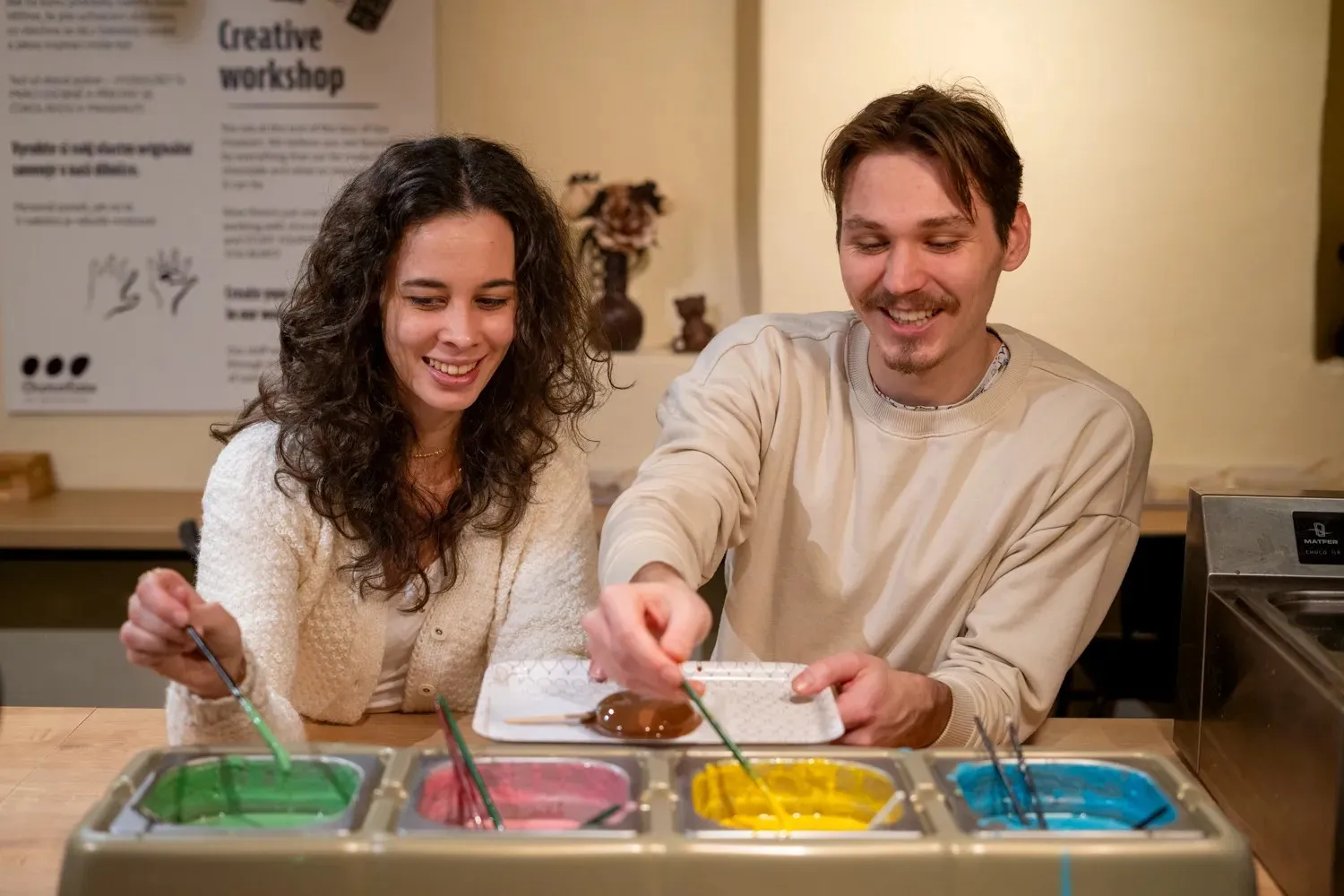 Twee lachende mensen die deelnemen aan een chocoladekunstworkshop met kleurrijke chocoladedips