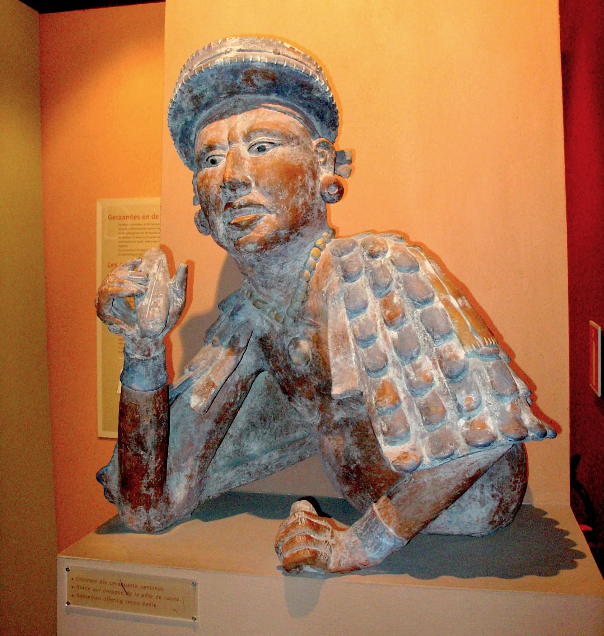 Escultura de cerámica pintada de un noble mesoamericano reclinado, con tocado y armadura de escamas, expuesta sobre un pedestal blanco contra una pared naranja de museo con un cartel informativo.