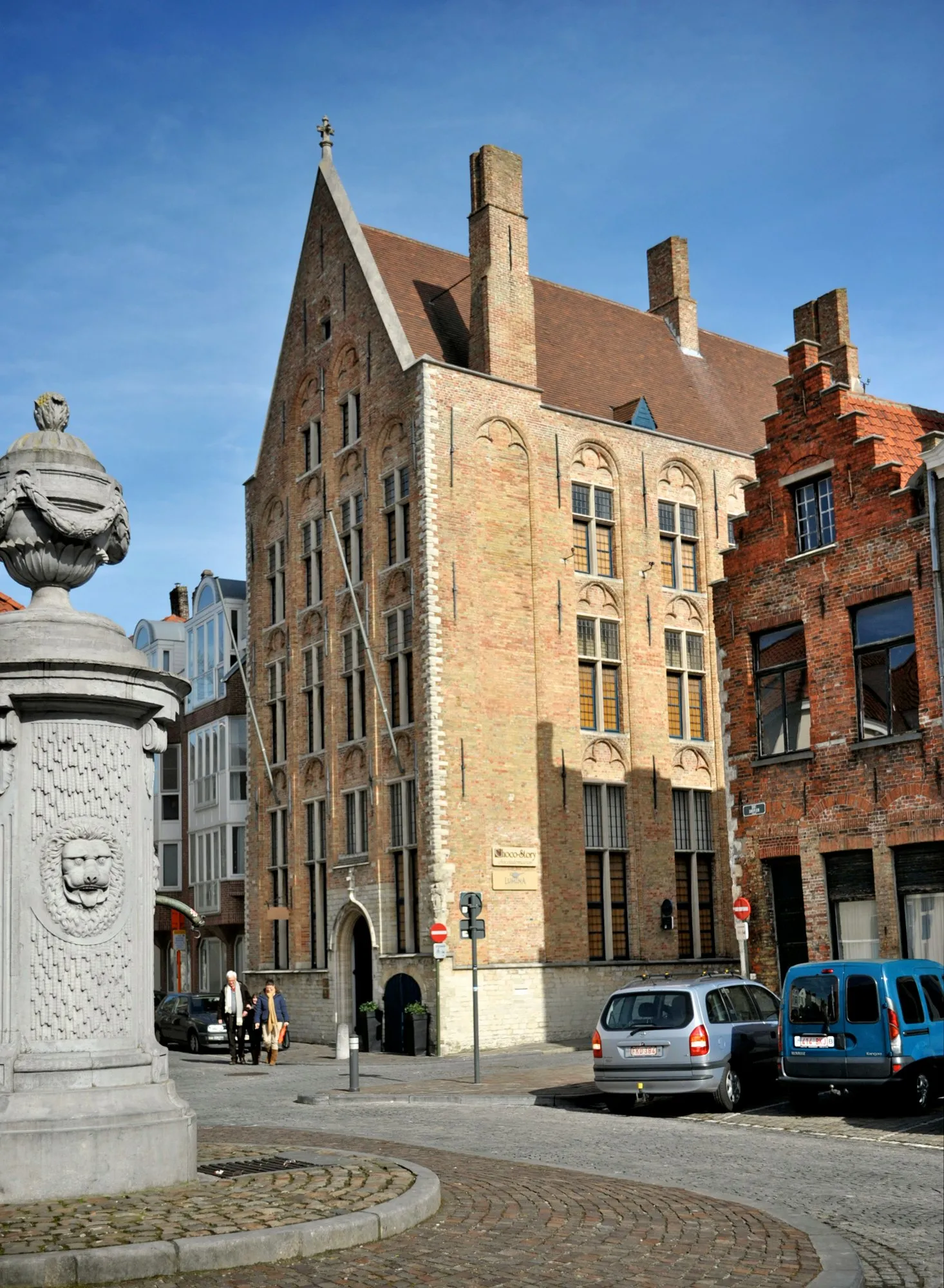 Histórico edificio de ladrillo con gablete escalonado en Brujas, con una fuente de piedra tallada en primer plano en una plaza adoquinada bajo un cielo azul despejado