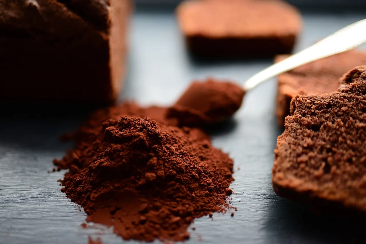 Un primer plano de un montón de rico cacao en polvo marrón sobre una superficie oscura, con una cuchara y trozos de pastel de chocolate al fondo.