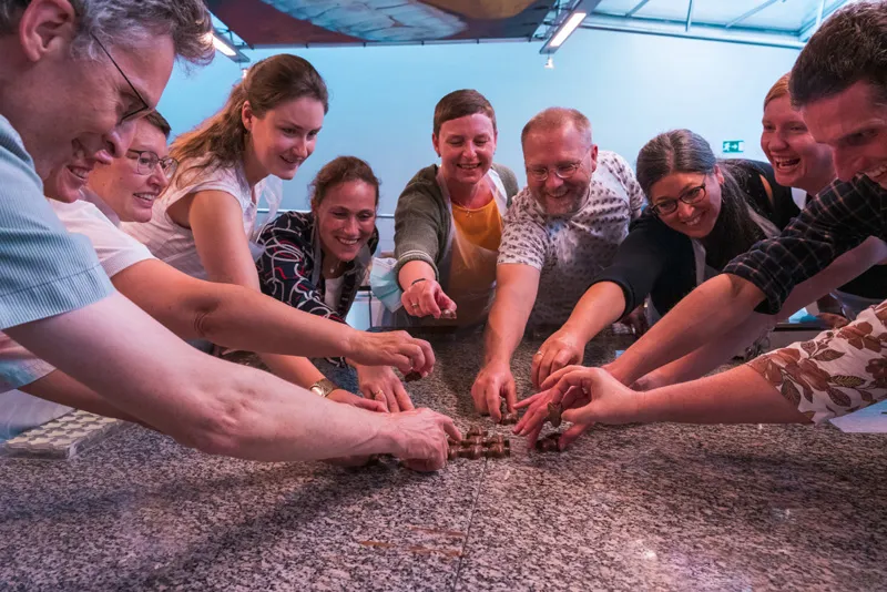 Grupo de personas sonrientes reunidas alrededor de una mesa de granito, extendiendo sus manos hacia chocolate o granos de cacao durante una demostración de taller.