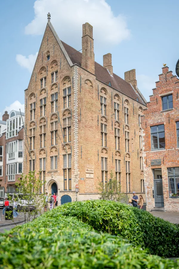Un histórico edificio de ladrillo de varias plantas con hastial escalonado y ventanas en arco, visto desde el otro lado de un seto verde con gente caminando cerca.