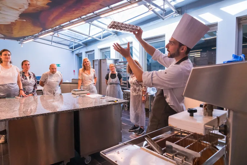 Un chef con un alto gorro blanco sostiene un molde de chocolate mientras hace una demostración ante un grupo de participantes con delantal que están de pie alrededor de mesas de acero inoxidable en un taller de cocina.