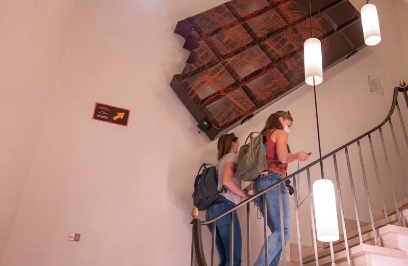 Dos visitantes subiendo una escalera blanca bajo una gran decoración de pared en forma de barra de chocolate y lámparas colgantes cilíndricas dentro de Choco-Story Brujas