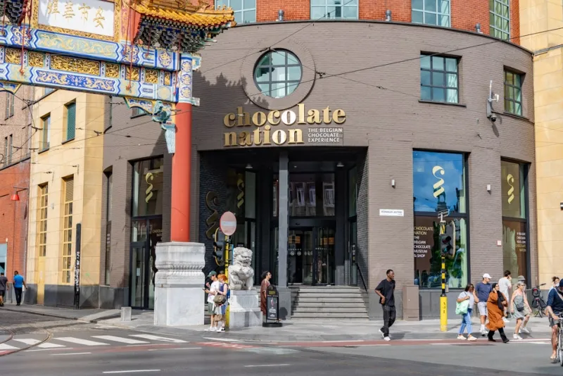 El edificio del museo Chocolate Nation con su fachada oscura curvada, señalización dorada y escalones de entrada, flanqueado por un arco tradicional chino con columnas rojas y una estatua de león de piedra, con peatones en la calle.