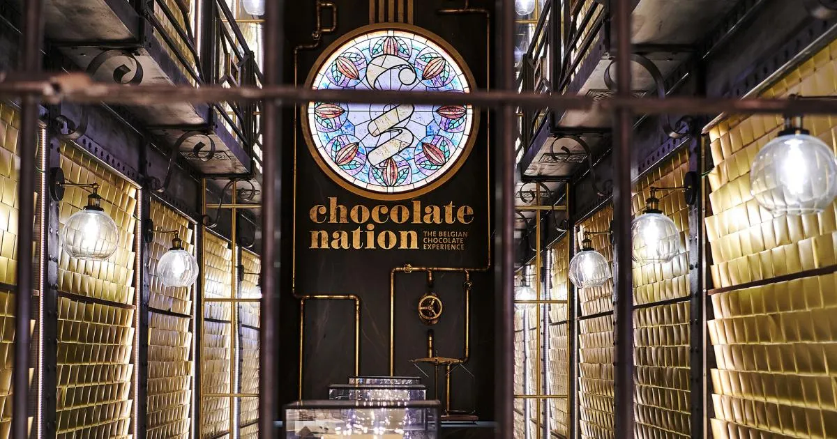 Pasillo interior del museo Chocolate Nation con paneles de pared en forma de barra de chocolate dorada, tuberías industriales, luces esféricas y un emblema circular de vidriera sobre el logotipo de Chocolate Nation en la pared del fondo
