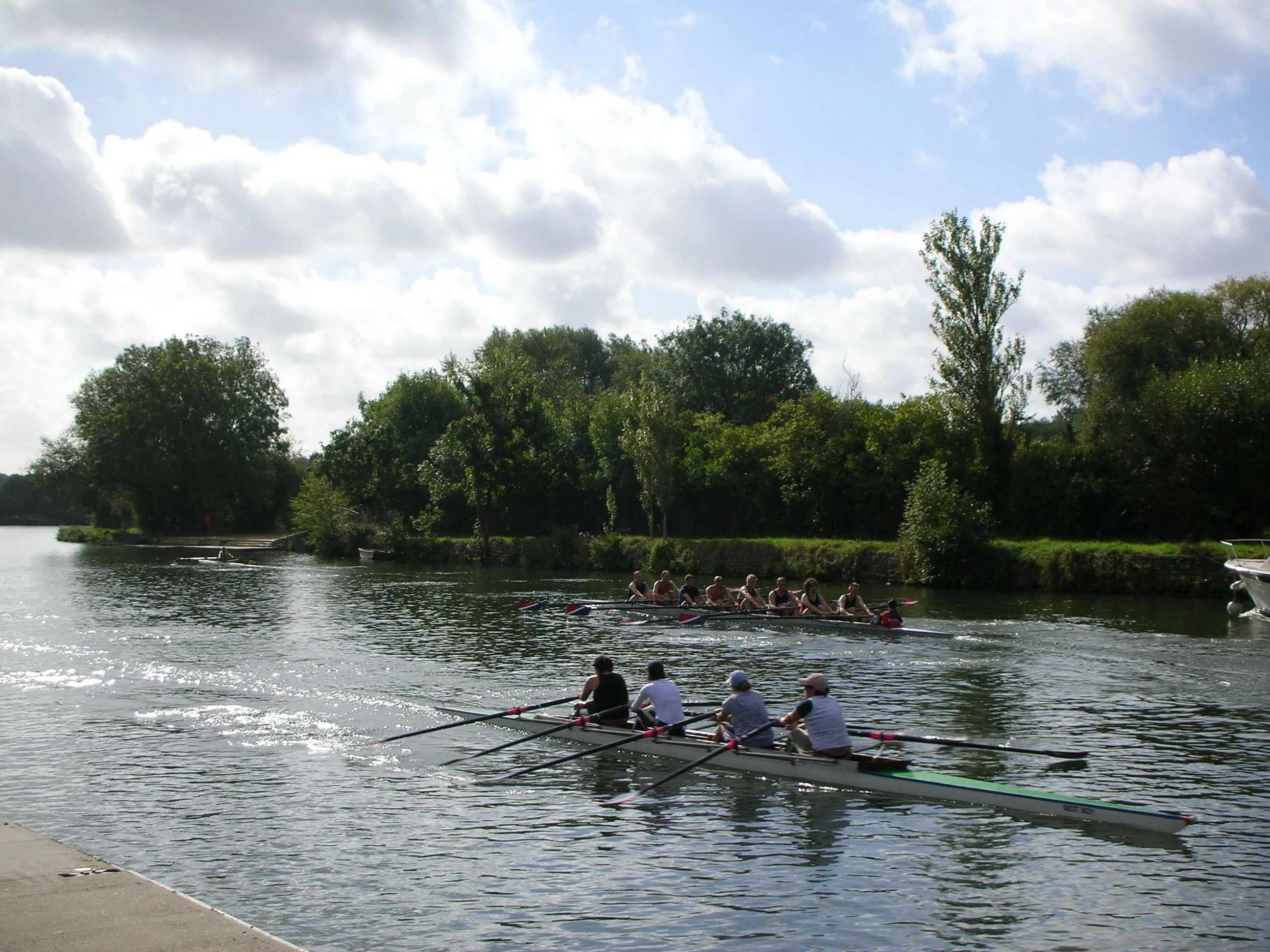 Rowers in a sculling boat on a river with trees and another rowing crew in the background under a cloudy sky.