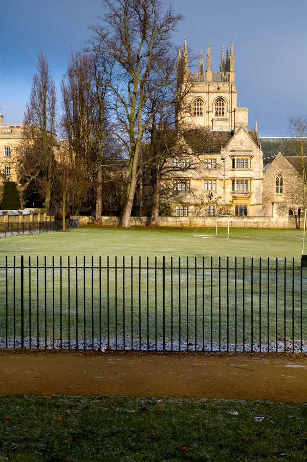 A frosty green sports field enclosed by a black iron fence, with bare trees and the historic stone tower of Christ Church College rising in the background under a blue sky.