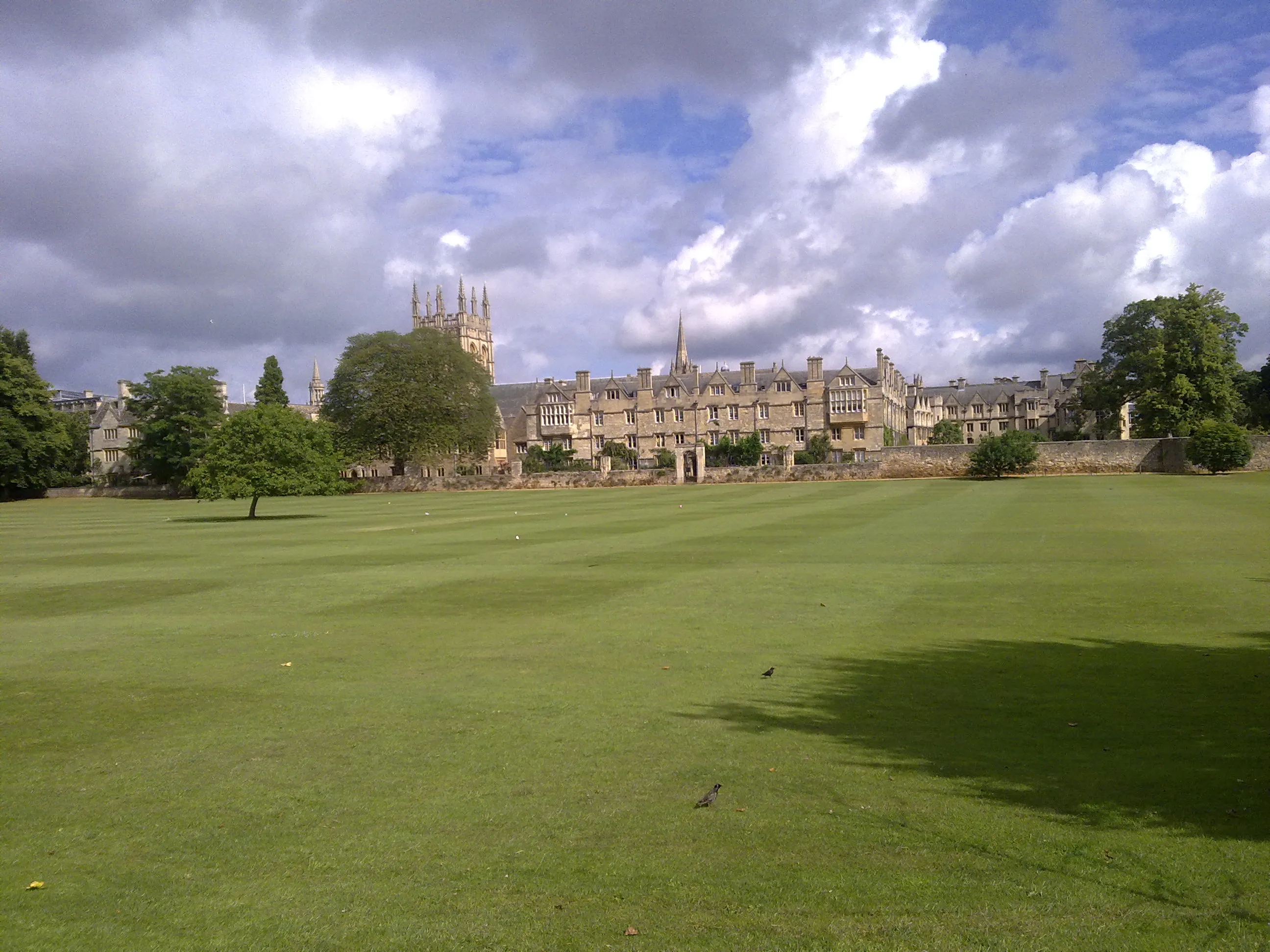 A wide green meadow stretches toward historic Oxford college buildings with a Gothic tower rising against a cloudy sky.