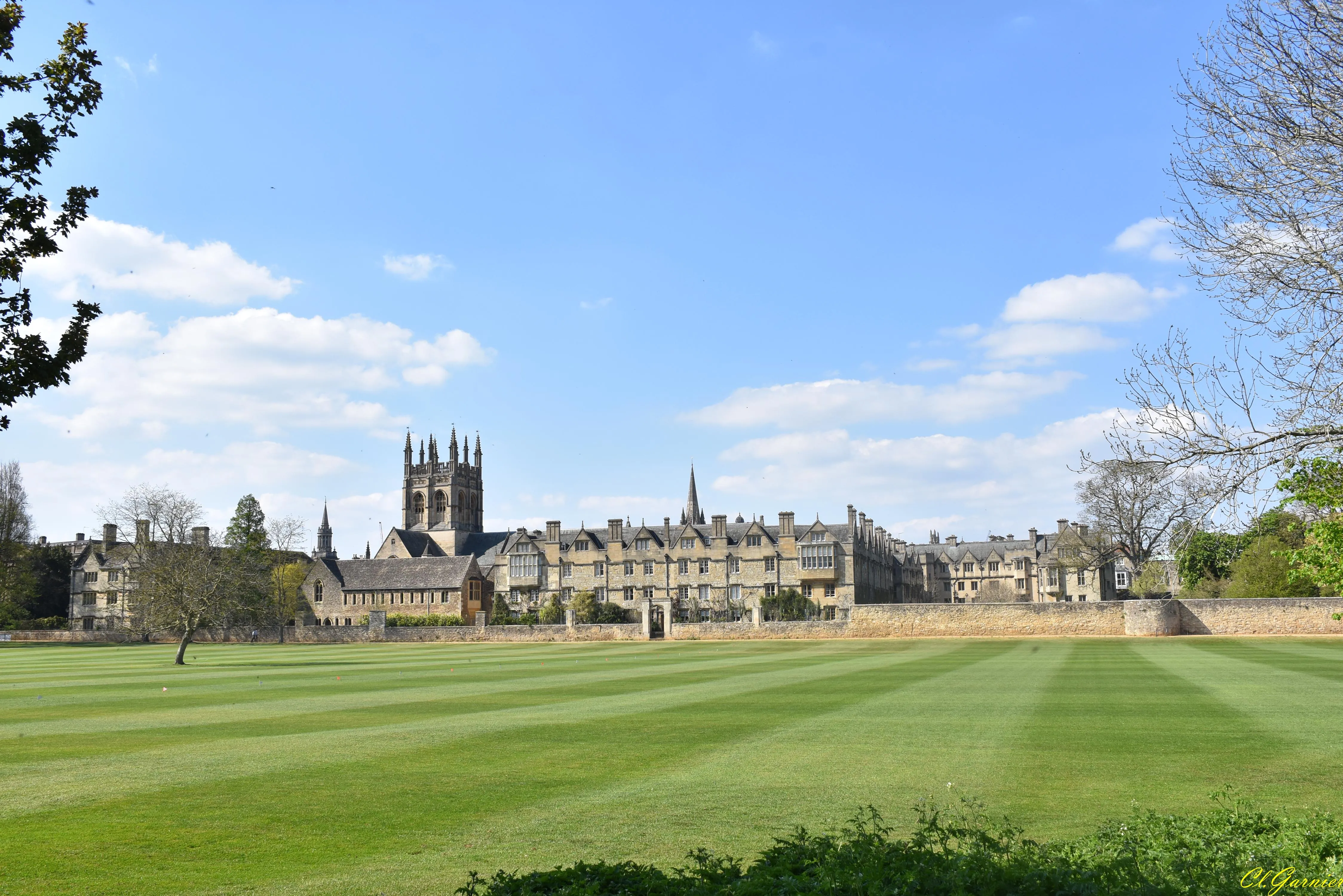Wide view of Christ Church college buildings with a Gothic tower across a large green mowed meadow under a blue sky with scattered clouds
