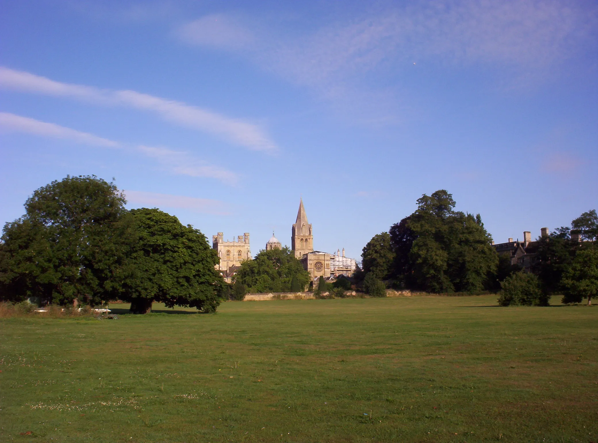 Wide green meadow with large leafy trees on either side and the Tom Tower spire of Christ Church Cathedral rising in the distance under a blue sky with wispy clouds.