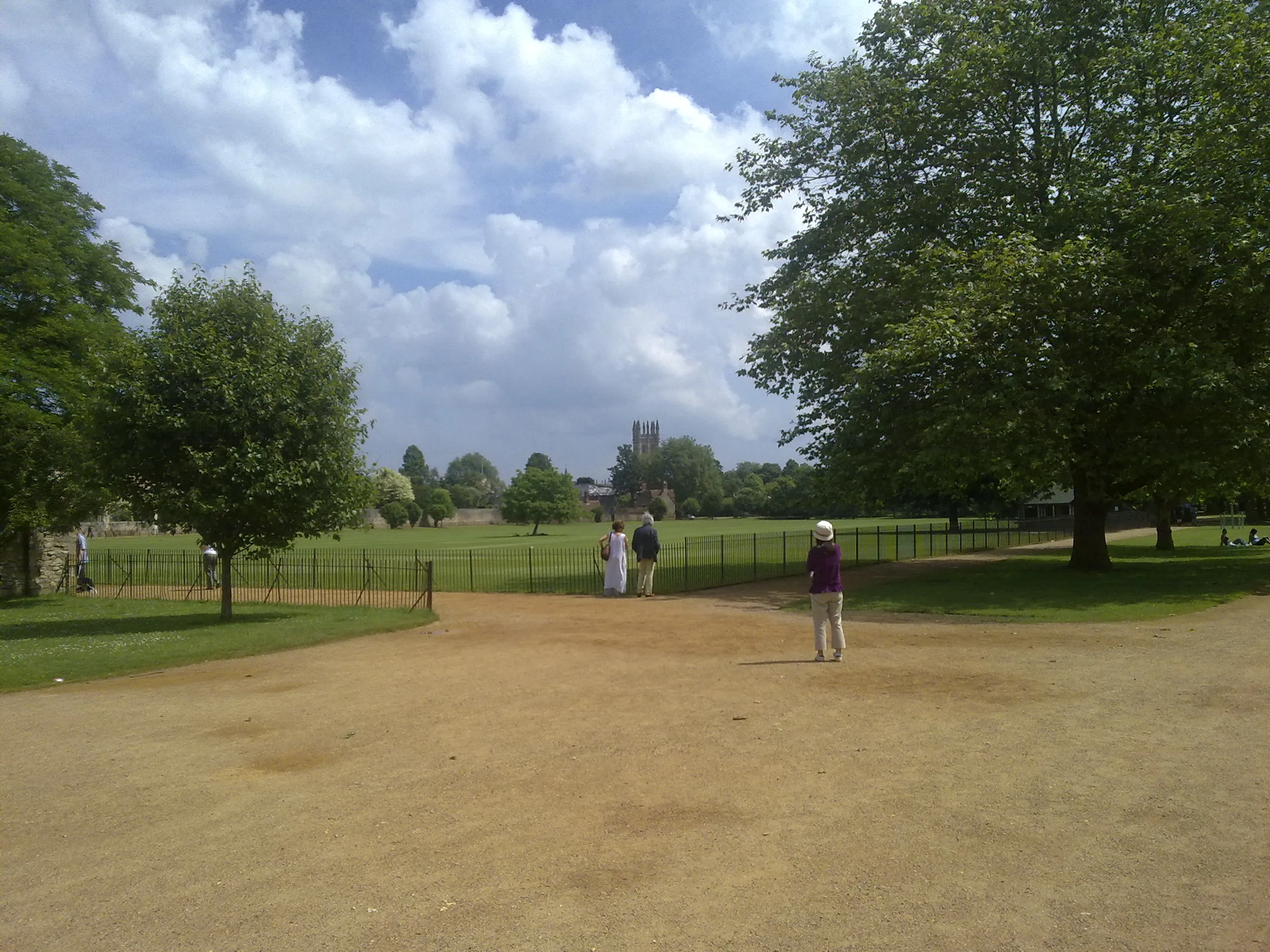 Wide gravel path through Christ Church Meadow with green trees, black iron fence, and Christ Church Cathedral tower visible in the distance under a blue sky with white clouds.