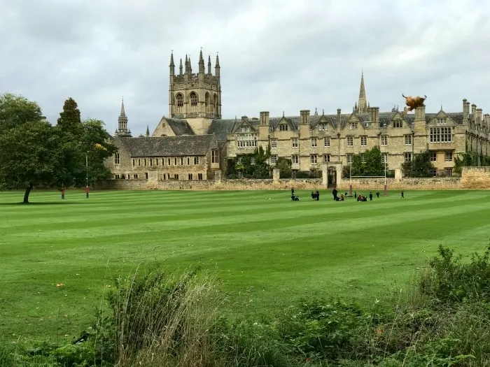 Wide green meadow with Christ Church's Tom Tower and historic college buildings in the background, with people walking across the grass under a cloudy sky