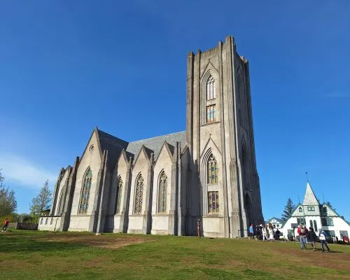 Catedral de piedra neogótica con una torre cuadrada alta y ventanas en arco apuntado, vista desde un césped bajo un cielo azul brillante con visitantes cerca de la entrada