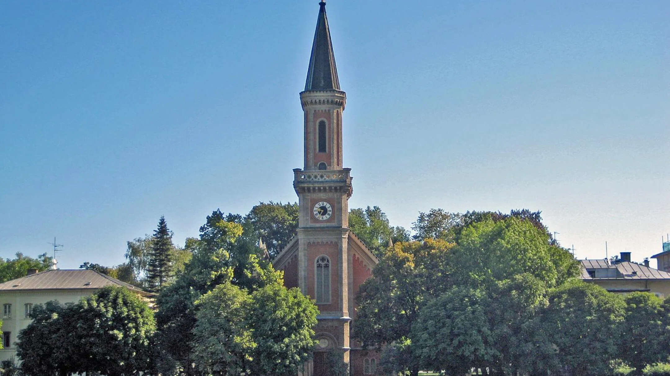 Alta torre de iglesia de ladrillo rojo con una aguja oscura y puntiaguda que se eleva sobre árboles verdes contra un cielo azul despejado