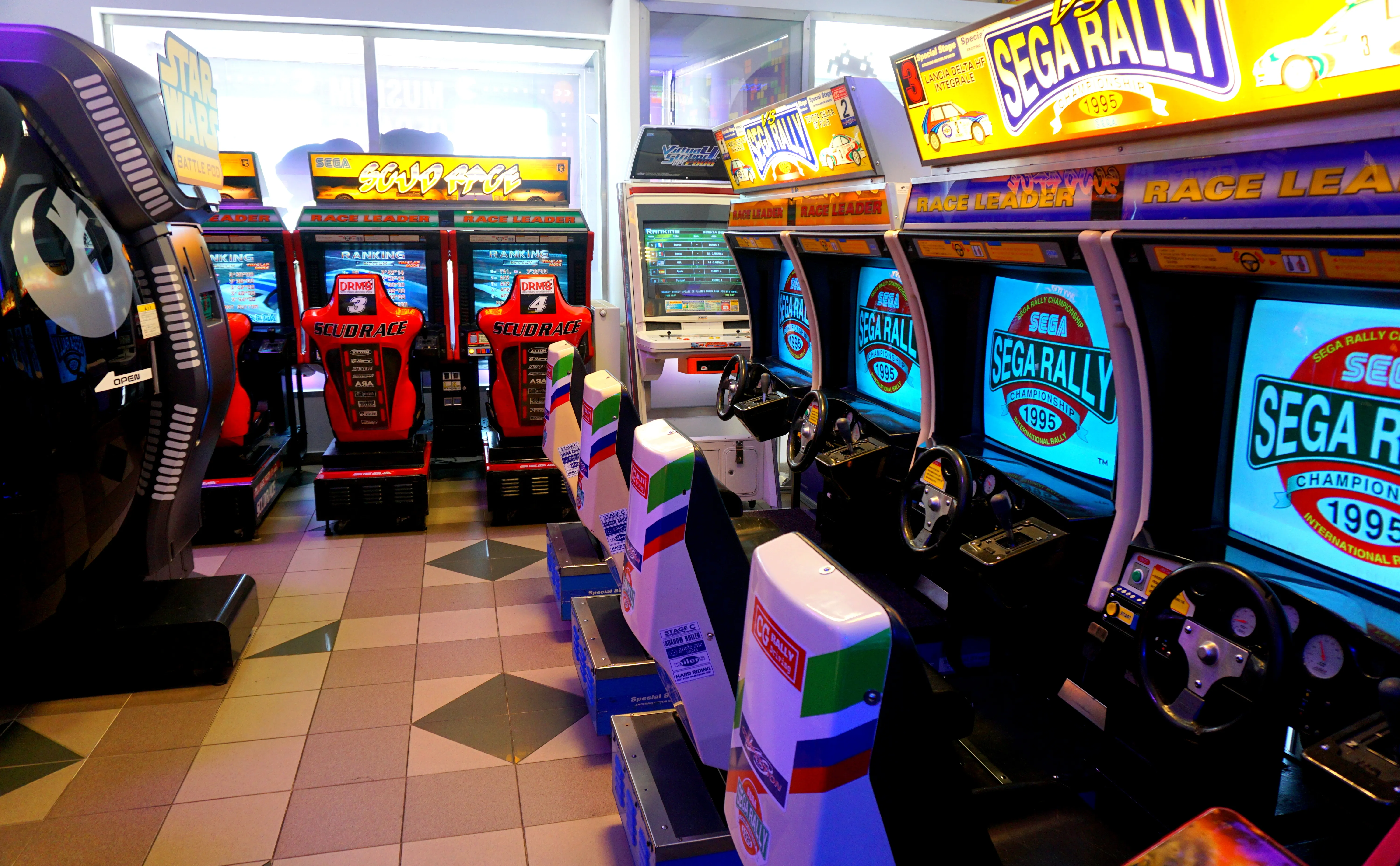 Arcade machines with Sega Rally branding and racing wheels in a museum gallery
