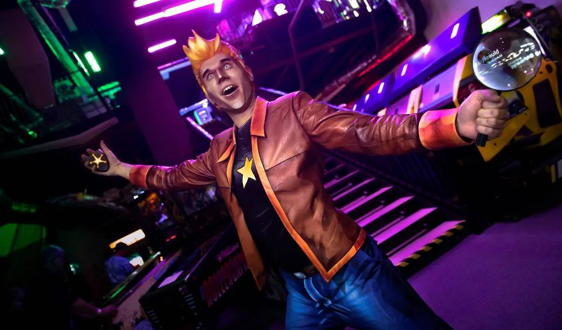 A life-sized figure of a video game character with spiky hair and a brown jacket stands with arms outstretched in a brightly lit arcade setting.
