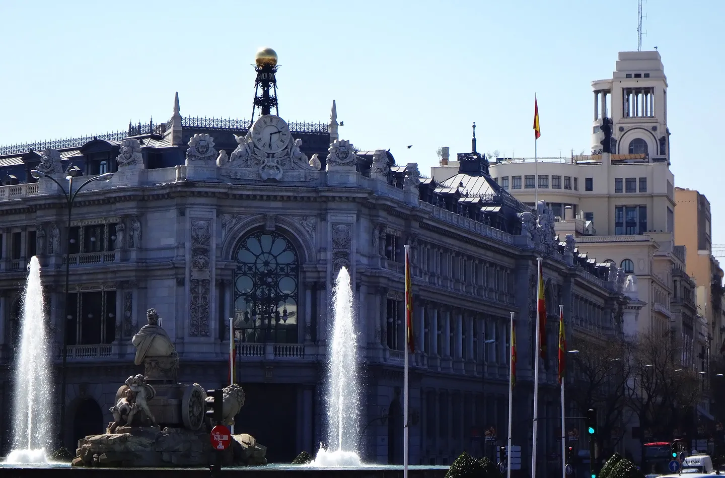 The Círculo de Bellas Artes building with ornate architecture, a central clock, and a golden sphere on the roof, with fountains in the foreground and Spanish flags on poles.