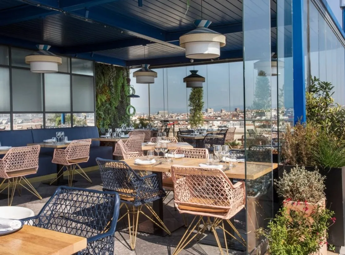 Rooftop terrace with wicker chairs and tables overlooking a cityscape through large glass windows