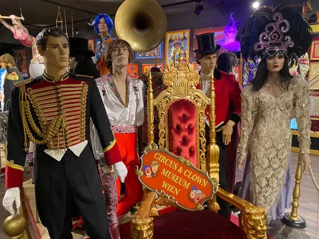Maniquíes con trajes de circo rodeando un trono dorado con un cartel del Circus- und Clownmuseum Wien