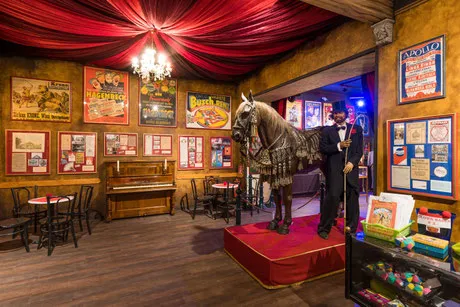 Interior de un museo con techo cubierto de tela roja, pósters enmarcados, un piano y una estatua de caballo sobre una plataforma roja