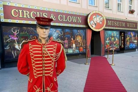 Maniquí con uniforme de circo rojo frente al Museo del Circo y el Payaso de Viena, con alfombra roja y murales coloridos en las ventanas
