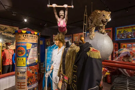 Maniquí en un trapecio, estatua de leopardo sobre un globo terráqueo y trajes de circo en una vitrina de museo