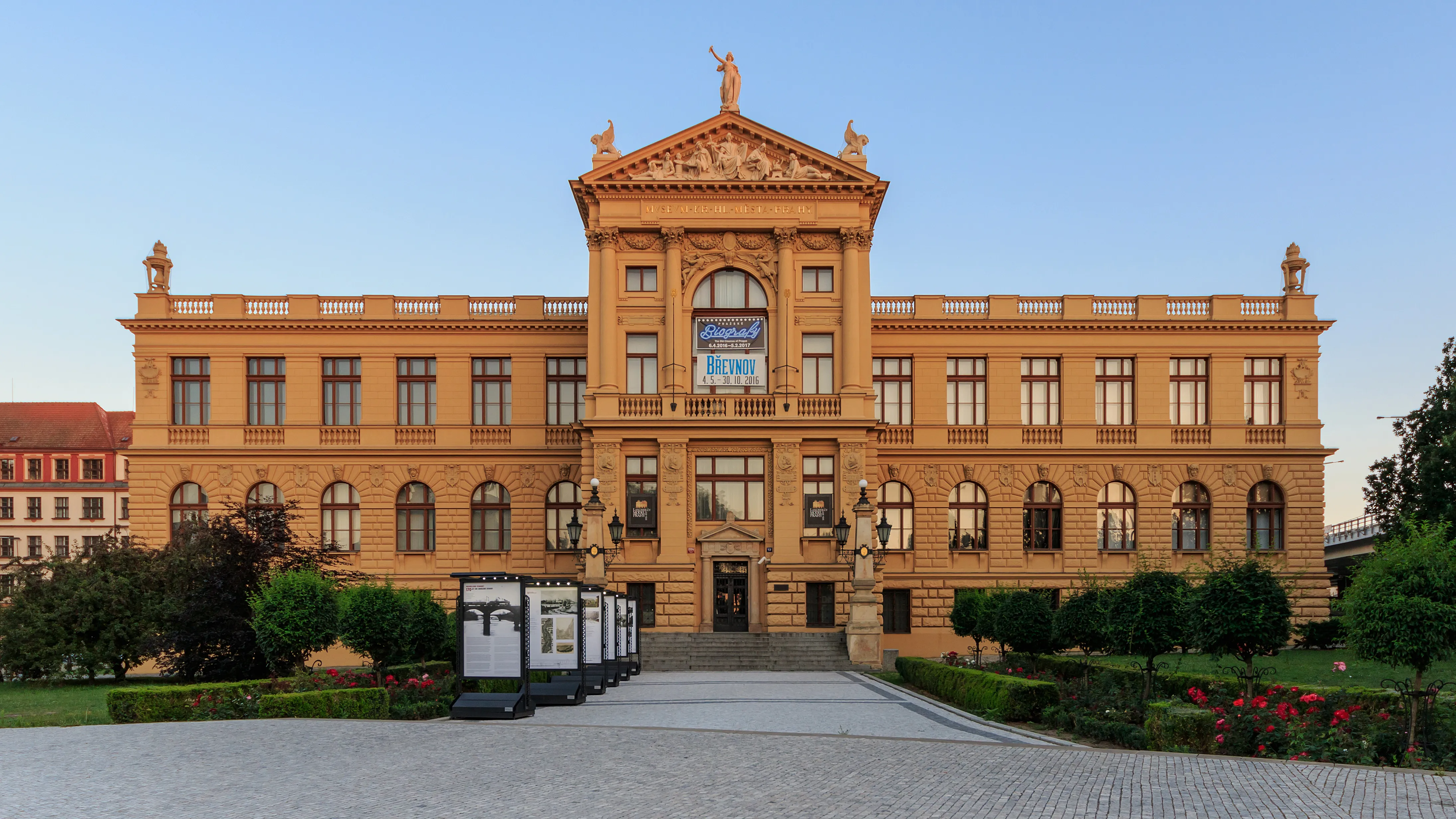 Neoclassical building with a central pediment and symmetrical facade, featuring large windows and a central entrance, surrounded by greenery and a paved courtyard.