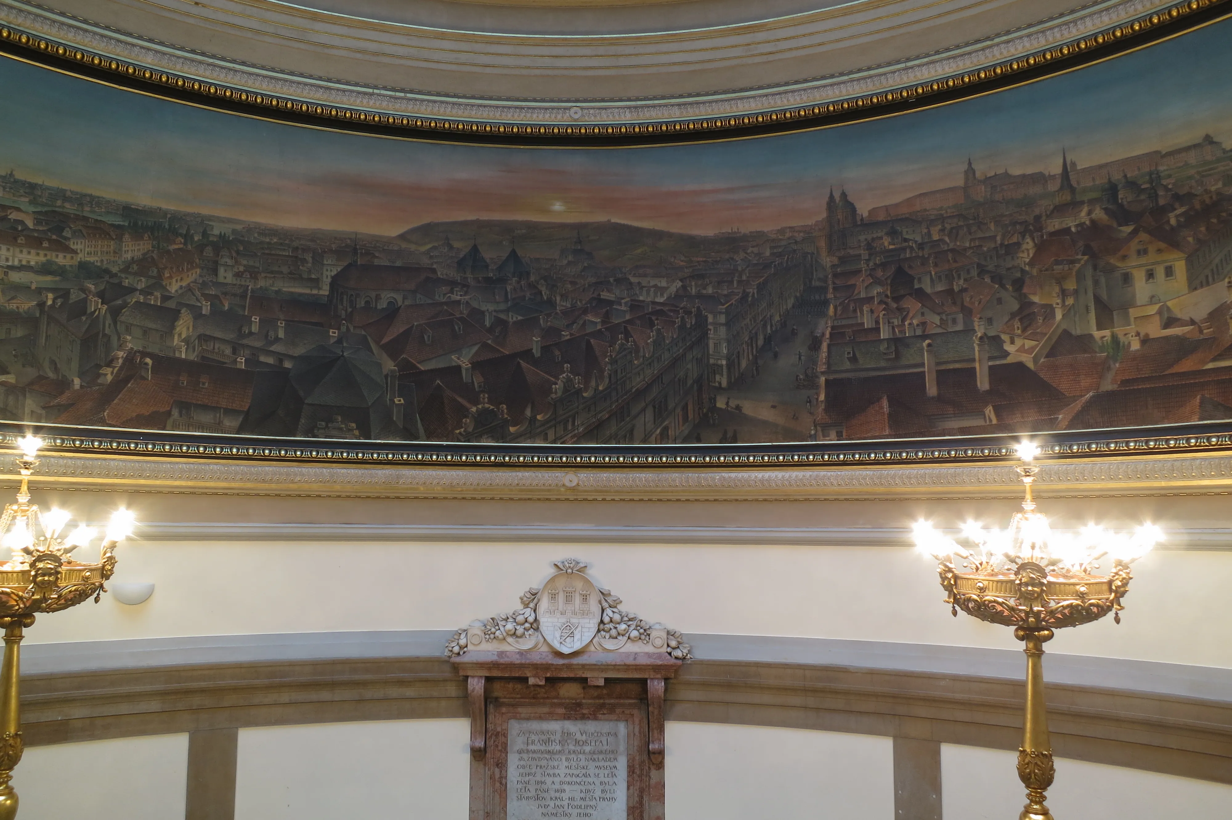 Interior hall with a large panoramic mural of Prague, ornate chandeliers, and a decorative plaque
