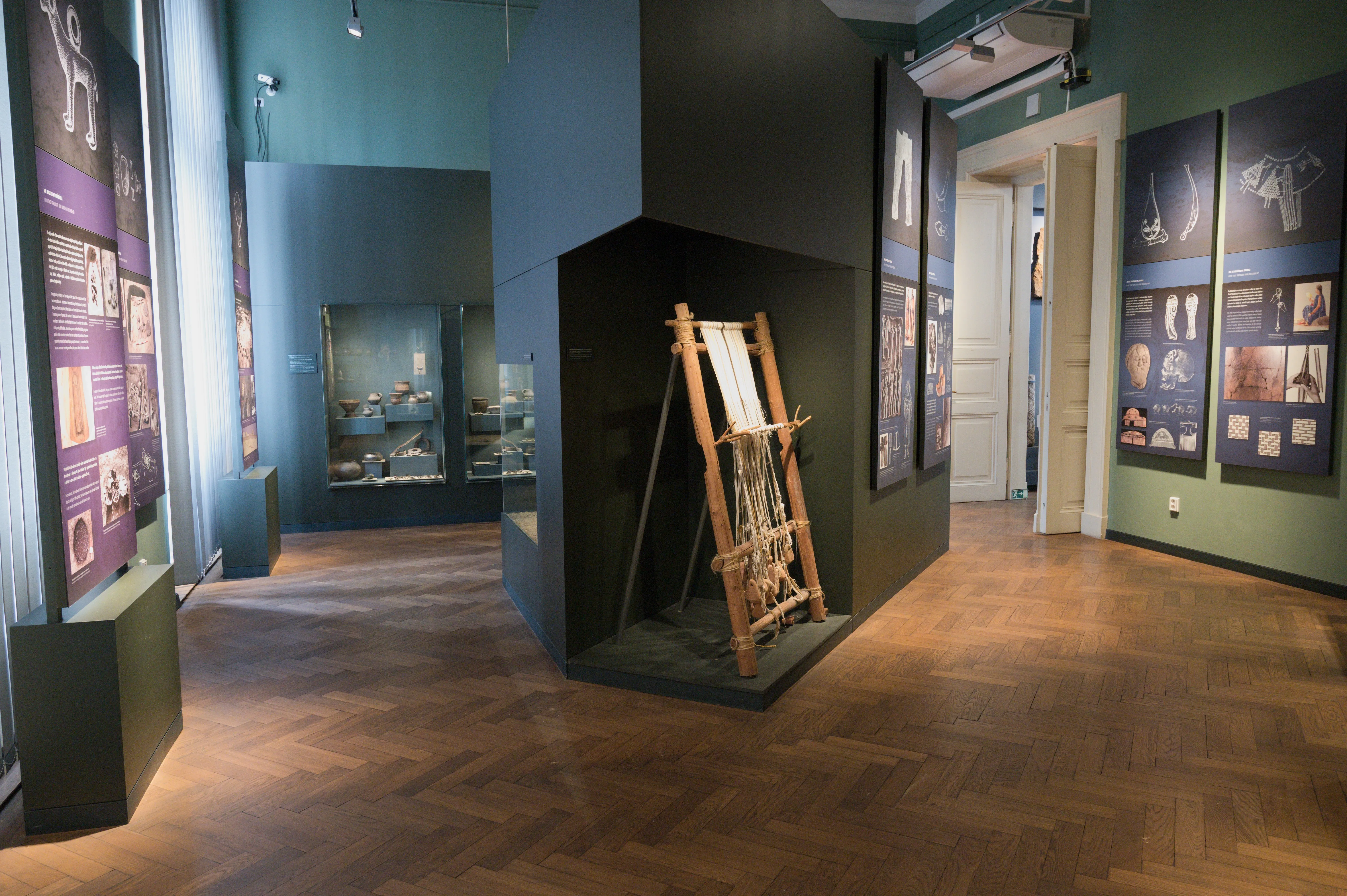 Interior of the City of Prague Museum with a wooden loom display, informational panels, and glass cases on dark green walls