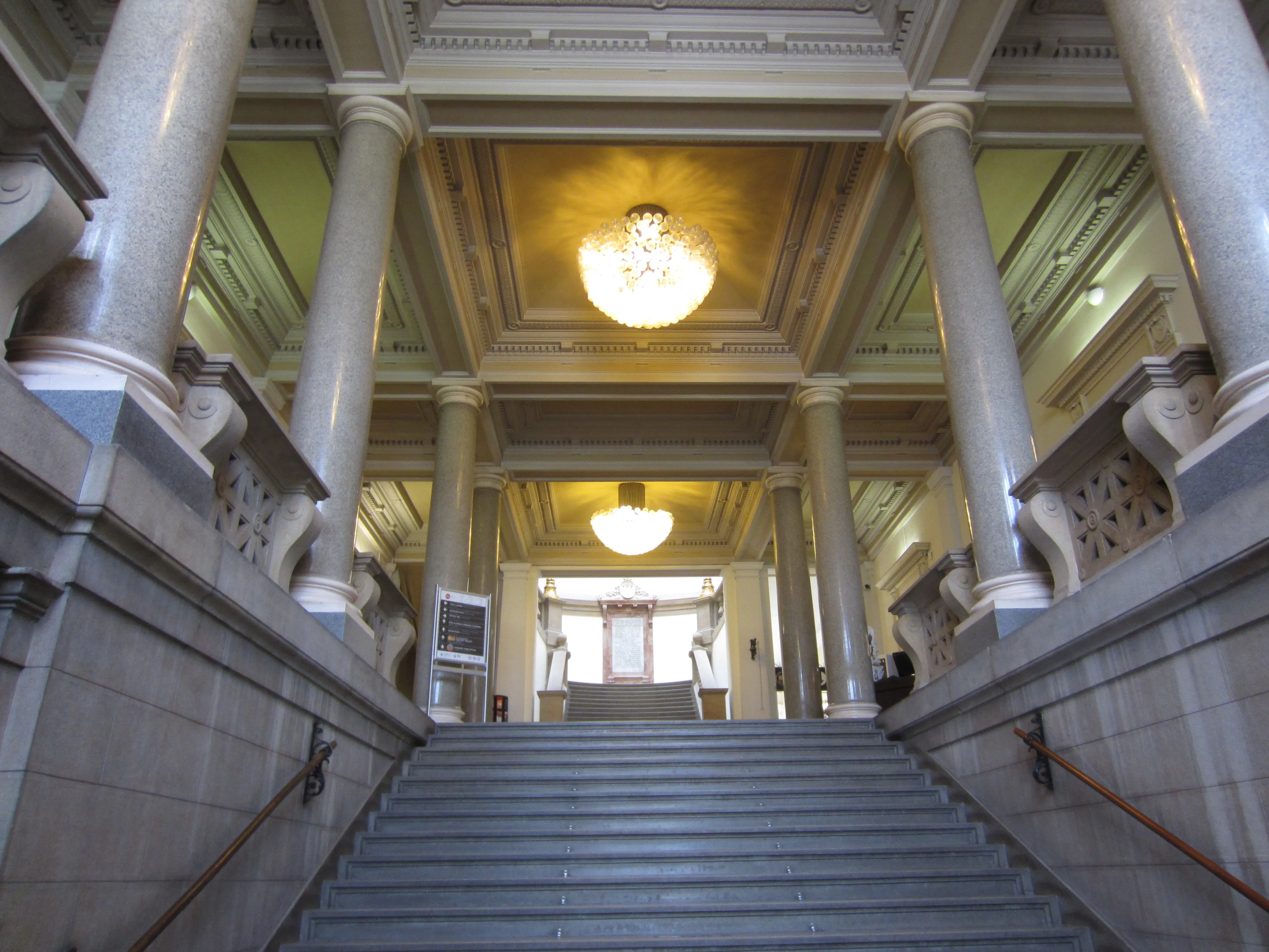 Grand staircase inside a museum with tall columns, ornate chandeliers, and a golden ceiling