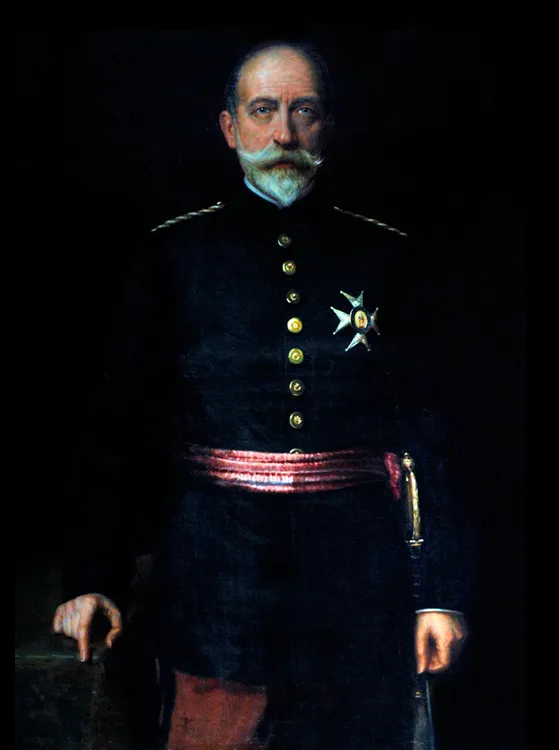 man in een donker militair uniform met gouden knopen en een rode sjerp