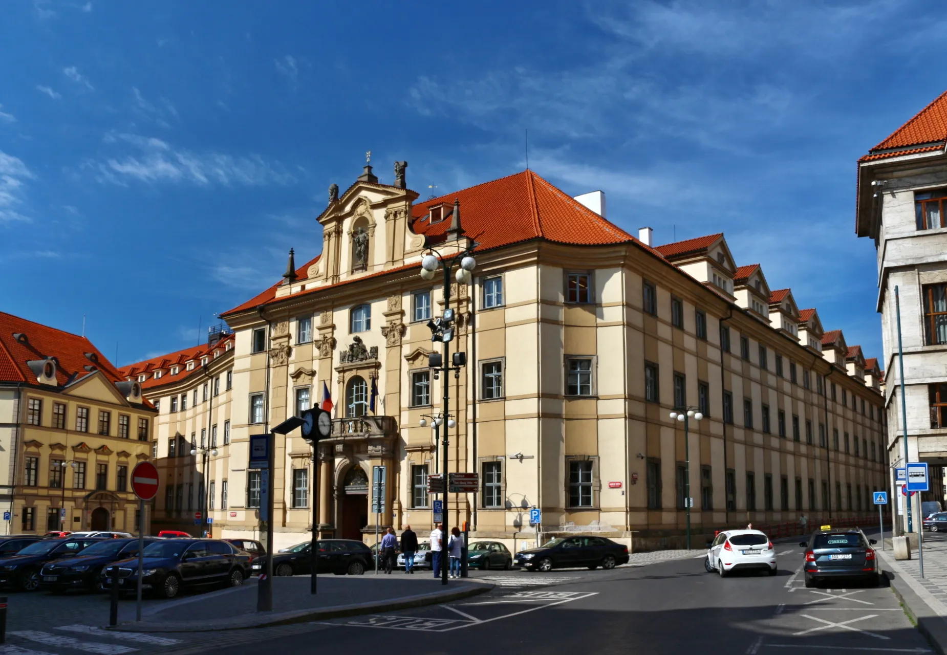 Un gran edificio histórico con fachada beige y tejado de tejas rojas, que presenta ornamentados detalles arquitectónicos barrocos y una entrada central, sobre un cielo azul con nubes dispersas.