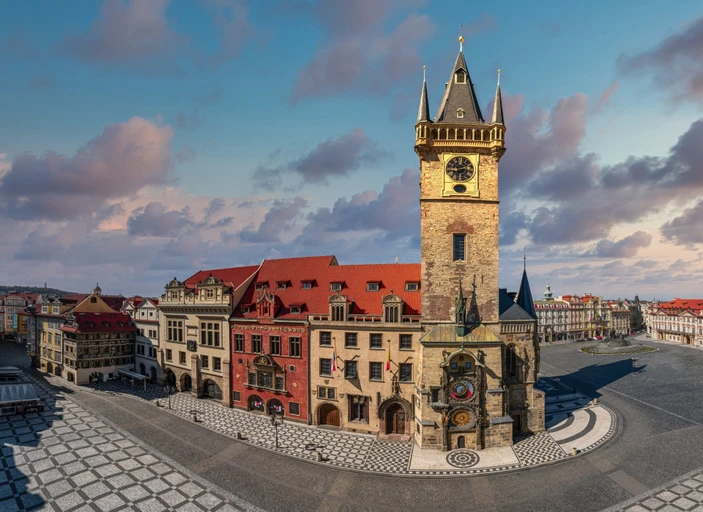 La Torre Astronómica del Clementinum con una esfera de reloj, rodeada de edificios históricos con tejados rojos y una plaza de piedra estampada bajo un cielo azul con nubes.