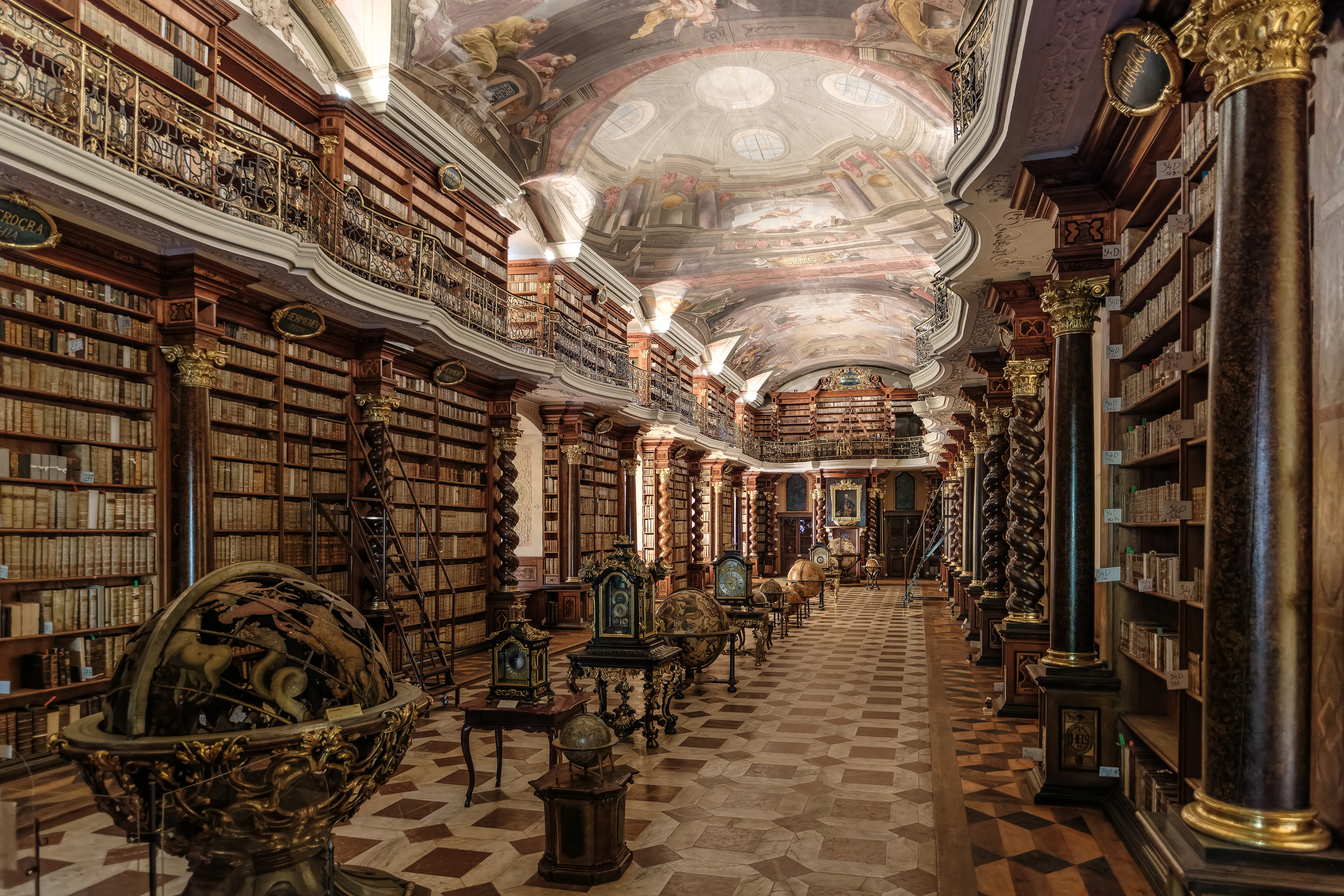 Largo salón de la Biblioteca Barroca del Clementinum con estanterías de suelo a techo, columnas ornamentadas, un techo pintado y un gran globo terráqueo antiguo en primer plano.