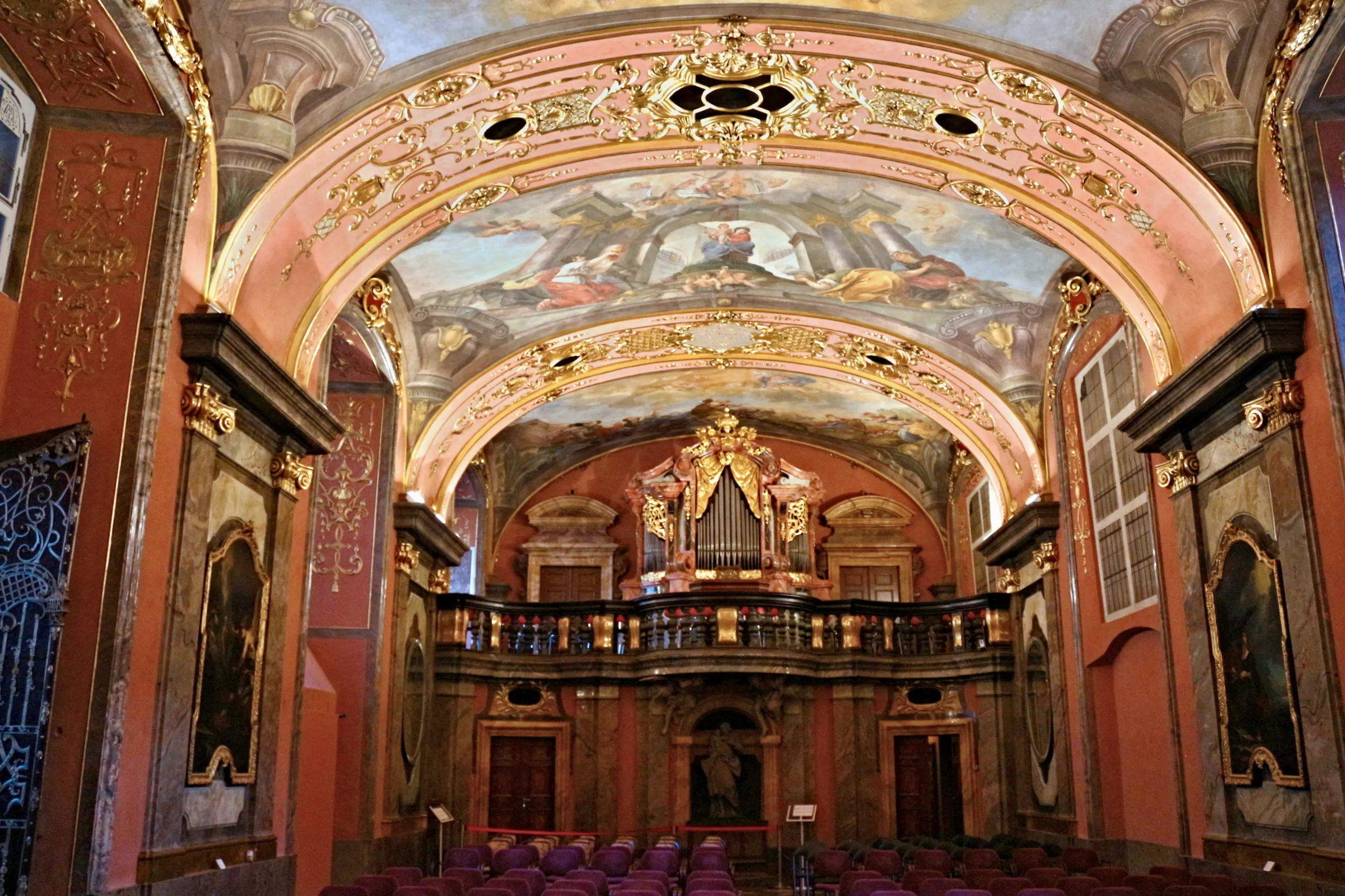 Interior barroco con un techo abovedado pintado con frescos, un órgano grande en un balcón y hileras de asientos morados.
