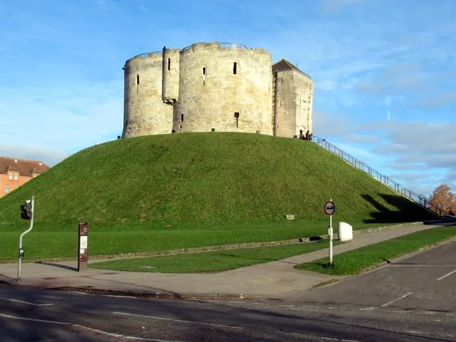 Clifford's Tower, kamienna wieża usytuowana na dużym, trawiastym wzgórzu, ze schodami prowadzącymi do wejścia i drogą na pierwszym planie pod błękitnym niebem.