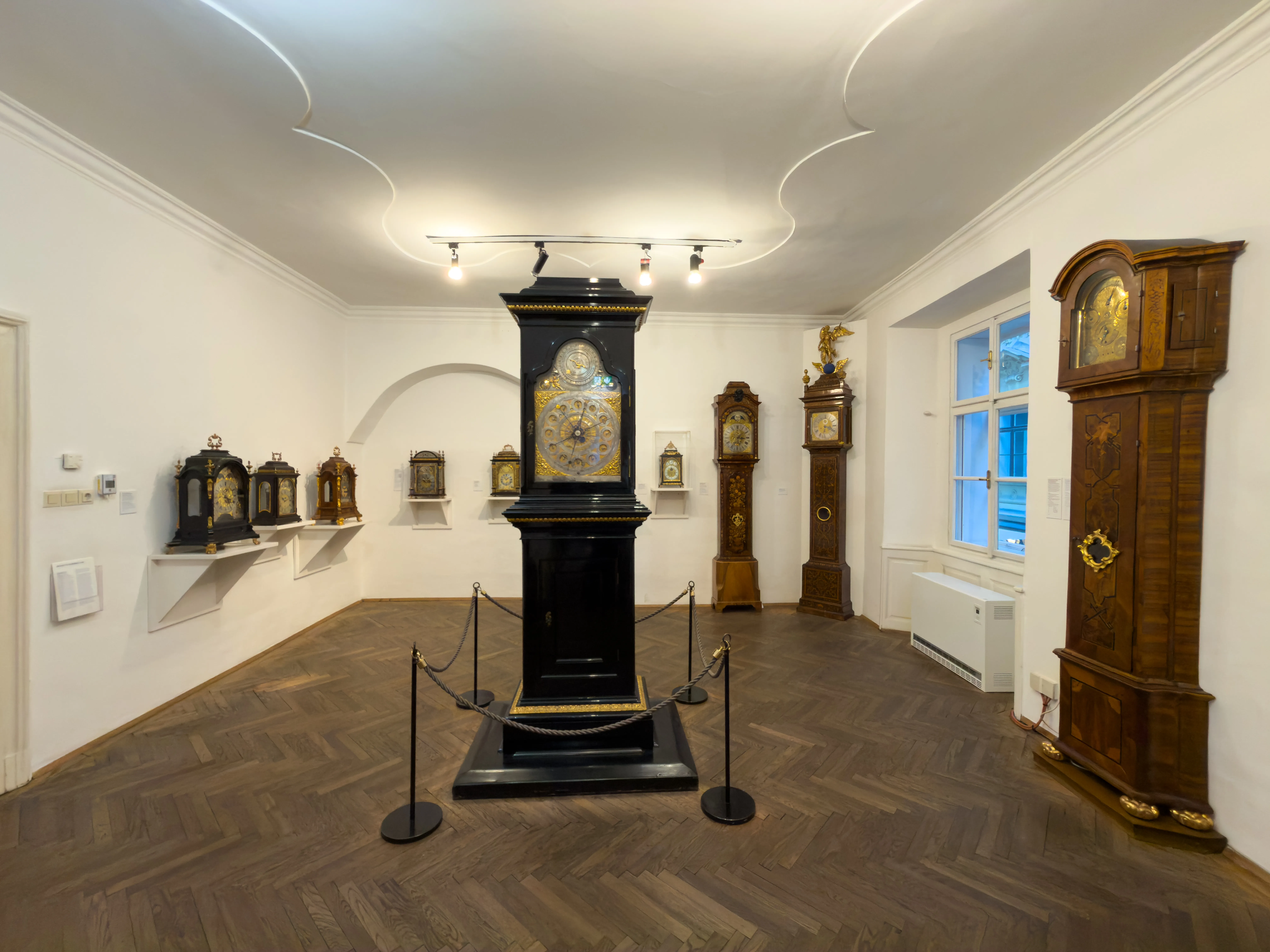 Interior de una sala de museo con una gran torre de reloj negra sobre un pedestal, rodeada de otros relojes en paredes y suelos.