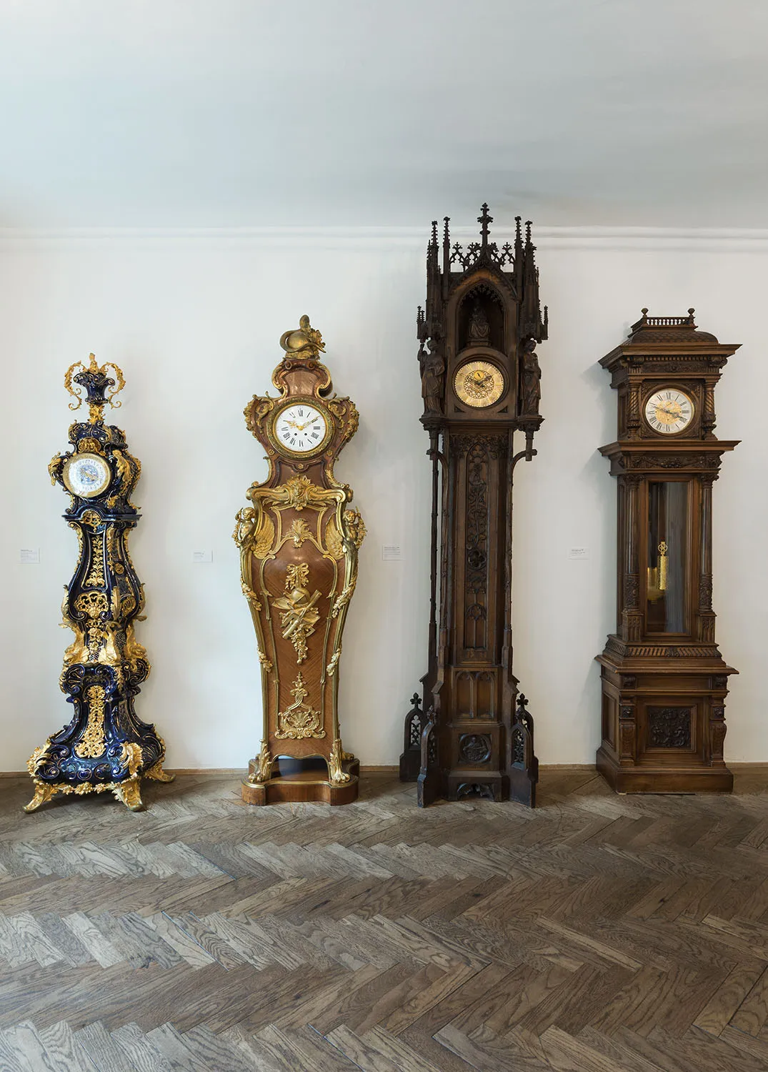 Cuatro altos y ornamentados relojes de pie con intrincadas tallas y elementos decorativos formaban una fila contra una pared blanca