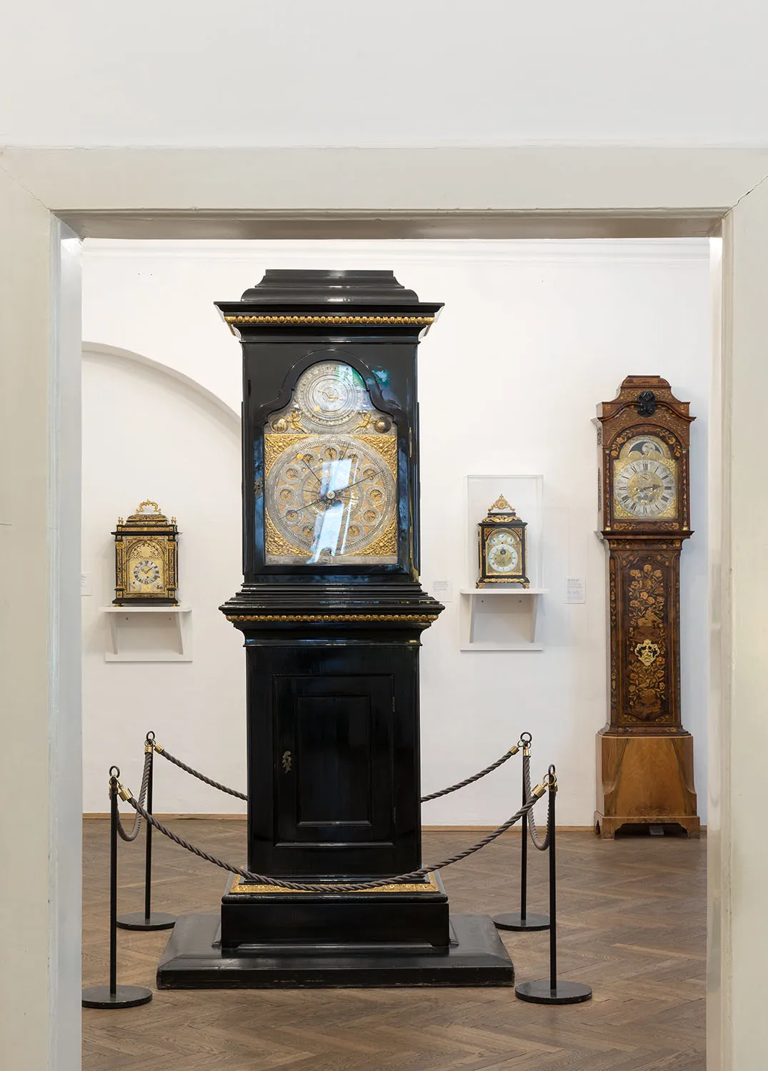 A large black and gold grandfather clock on display in a museum gallery, surrounded by other ornate clocks on white walls.