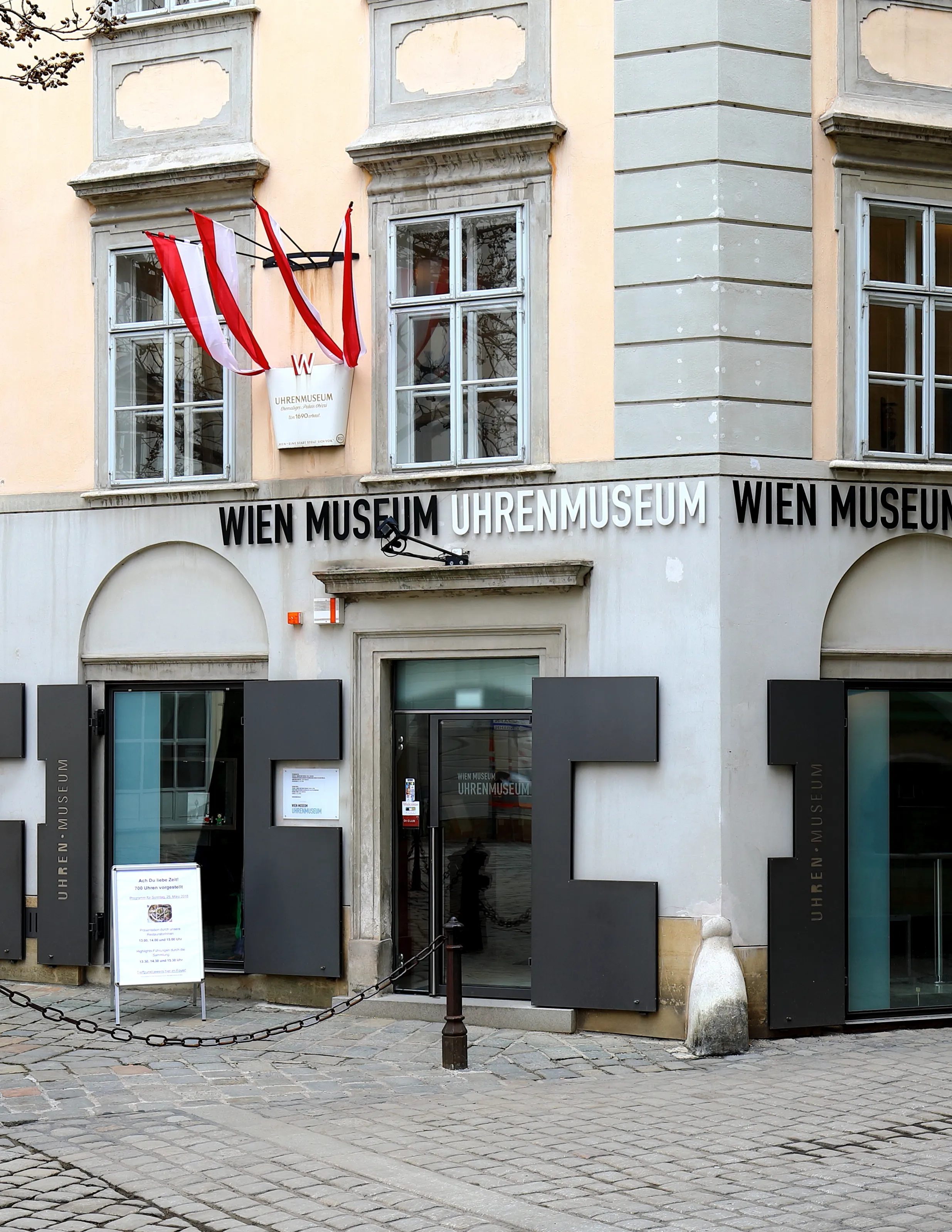 The exterior of the Wien Museum Uhrenmuseum with a sign above the entrance and Austrian flags on the building facade