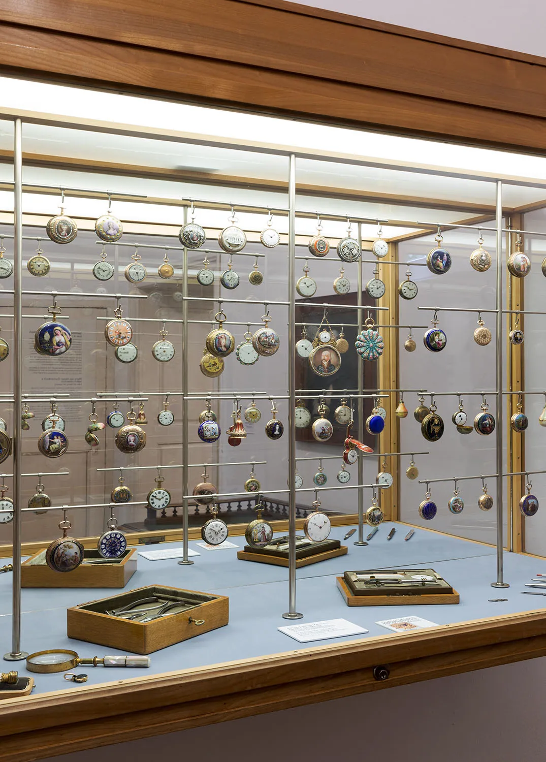 Una vitrina de cristal llena de numerosos relojes de bolsillo y de hora, algunos colgados de varillas metálicas y otros en cajas de madera sobre una superficie azul.