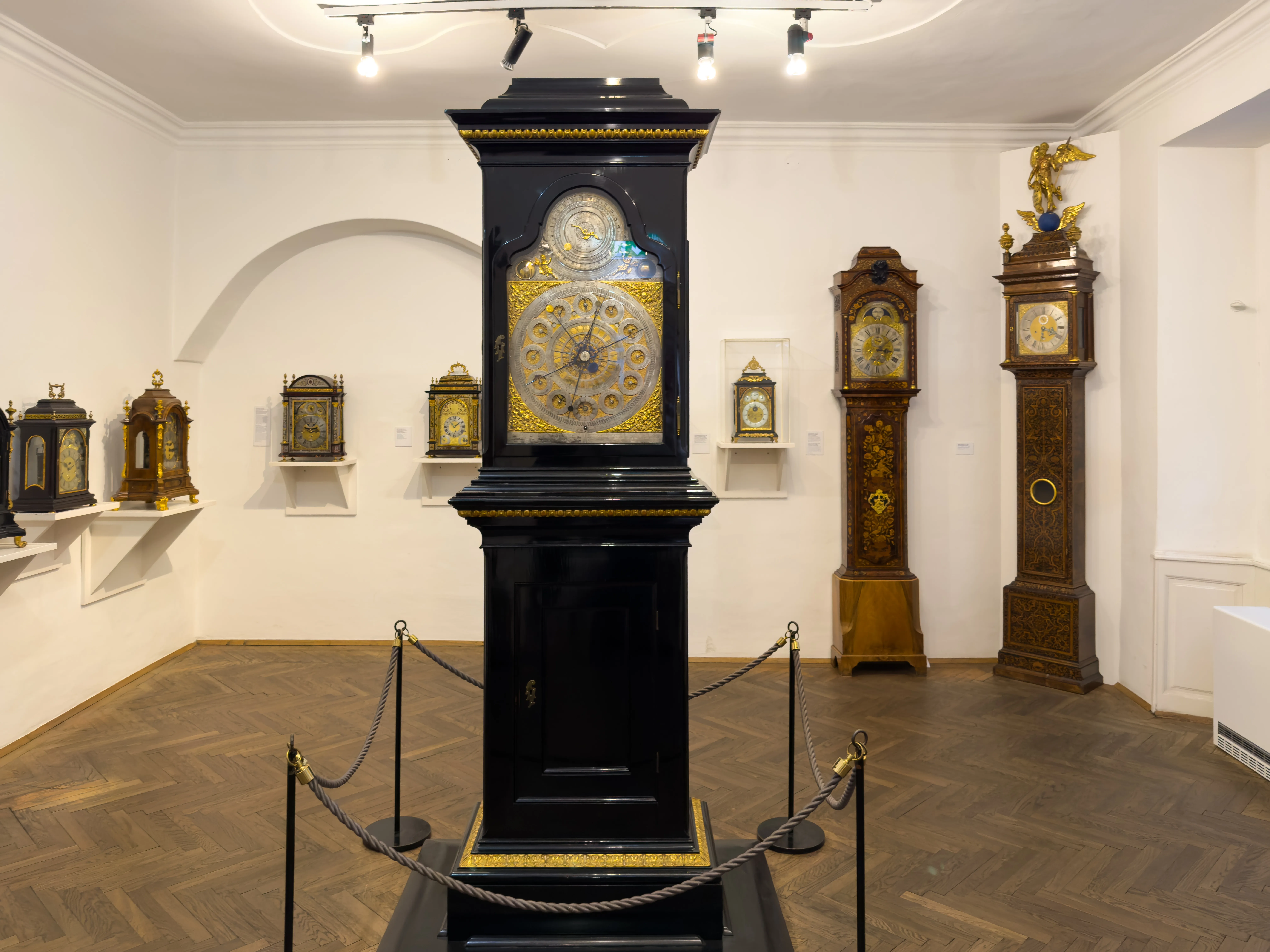 A large black and gold grandfather clock on display in a museum gallery, surrounded by other clocks on white walls and a wooden floor.